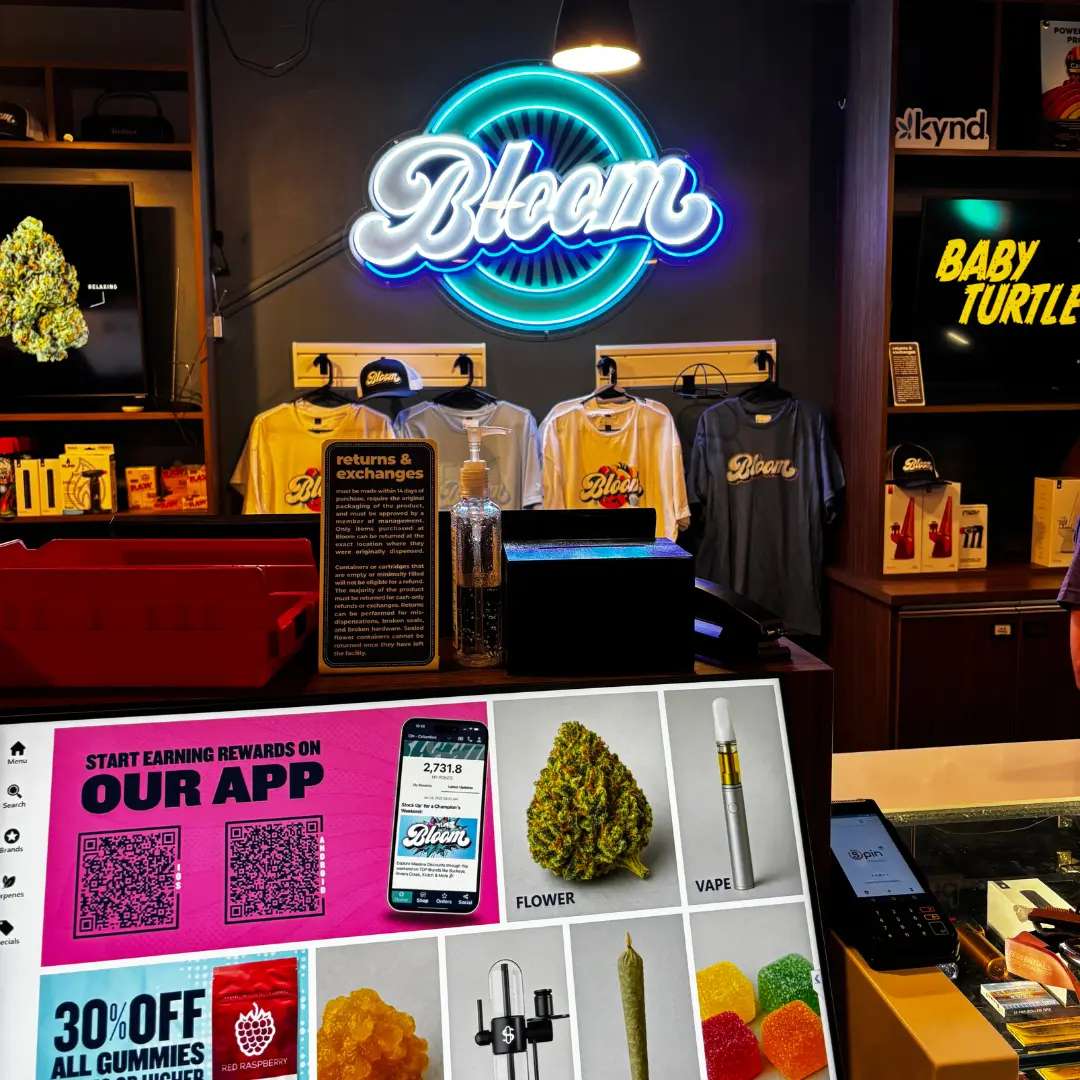 Bloom west columbus dispensary counter with "Bloom" neon sign, T-shirts, and a display showcasing vape pens, cannabis, and gummies. Promo for app rewards visible.