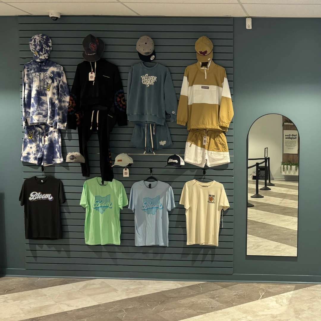 Bloom south columbus dispensary wall with clothing display on a teal wall with various shirts, shorts, and hats. There is a tall mirror on the right and a rope barrier in the background.