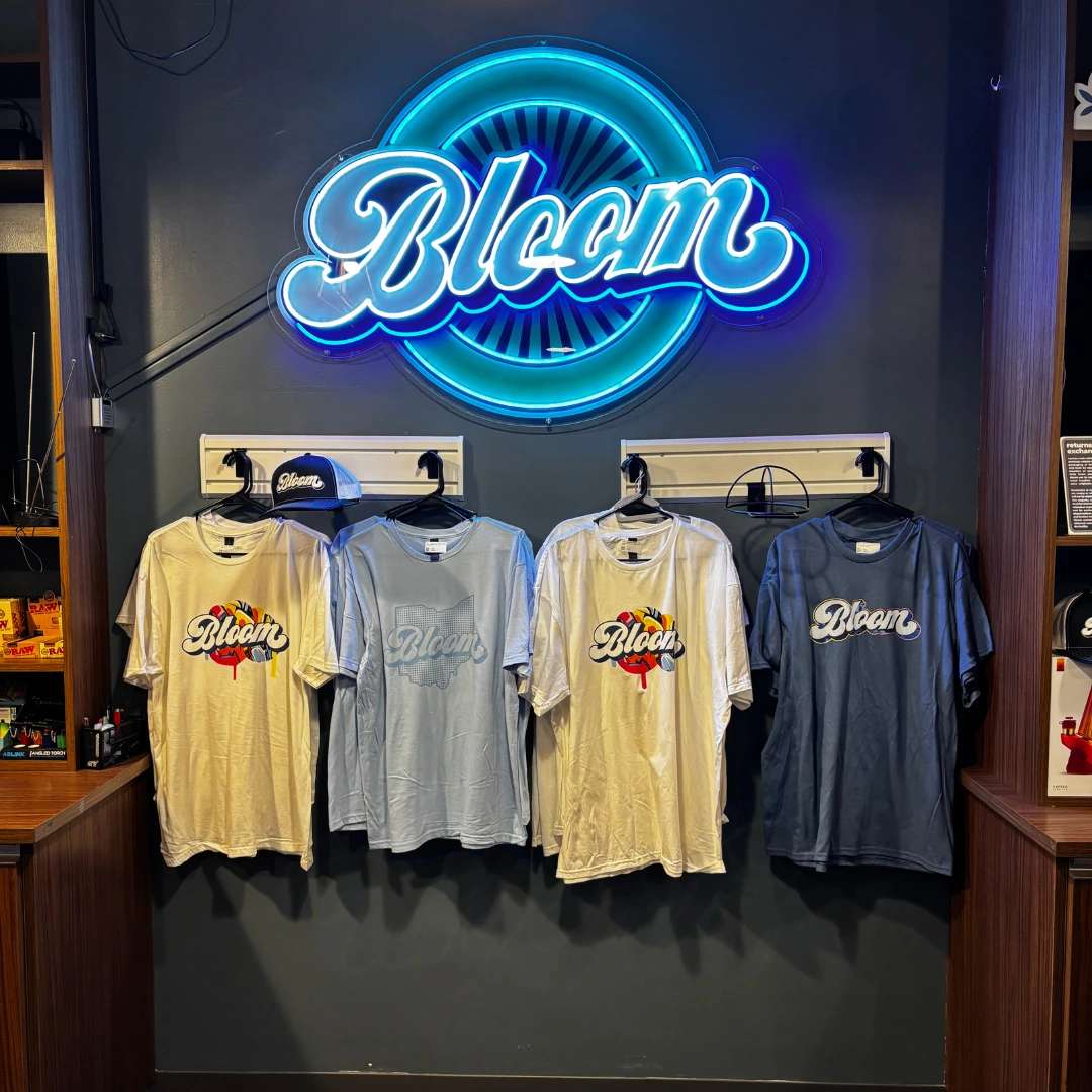 Bloom west columbus dispensary with four t-shirts with "Bloom" printed hang under a bright neon "Bloom" sign on a dark wall.