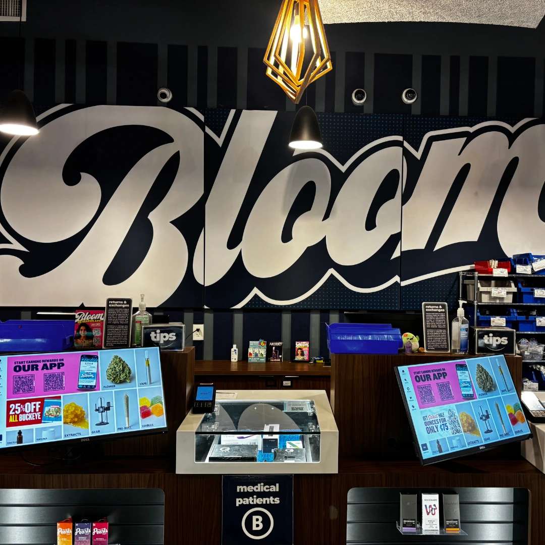 Bloom west columbus dispensary interior with "Bloom" on the wall, digital displays on counters, and shelves with various products. Modern lighting hangs from the ceiling.
