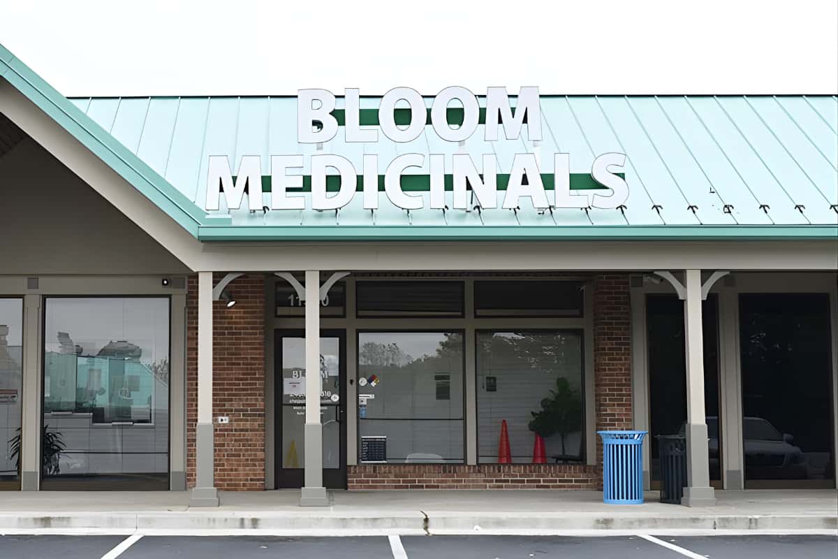 Storefront of Bloom Medicinals with large white sign on teal metal roof, brick facade, empty windows and a blue trash can.