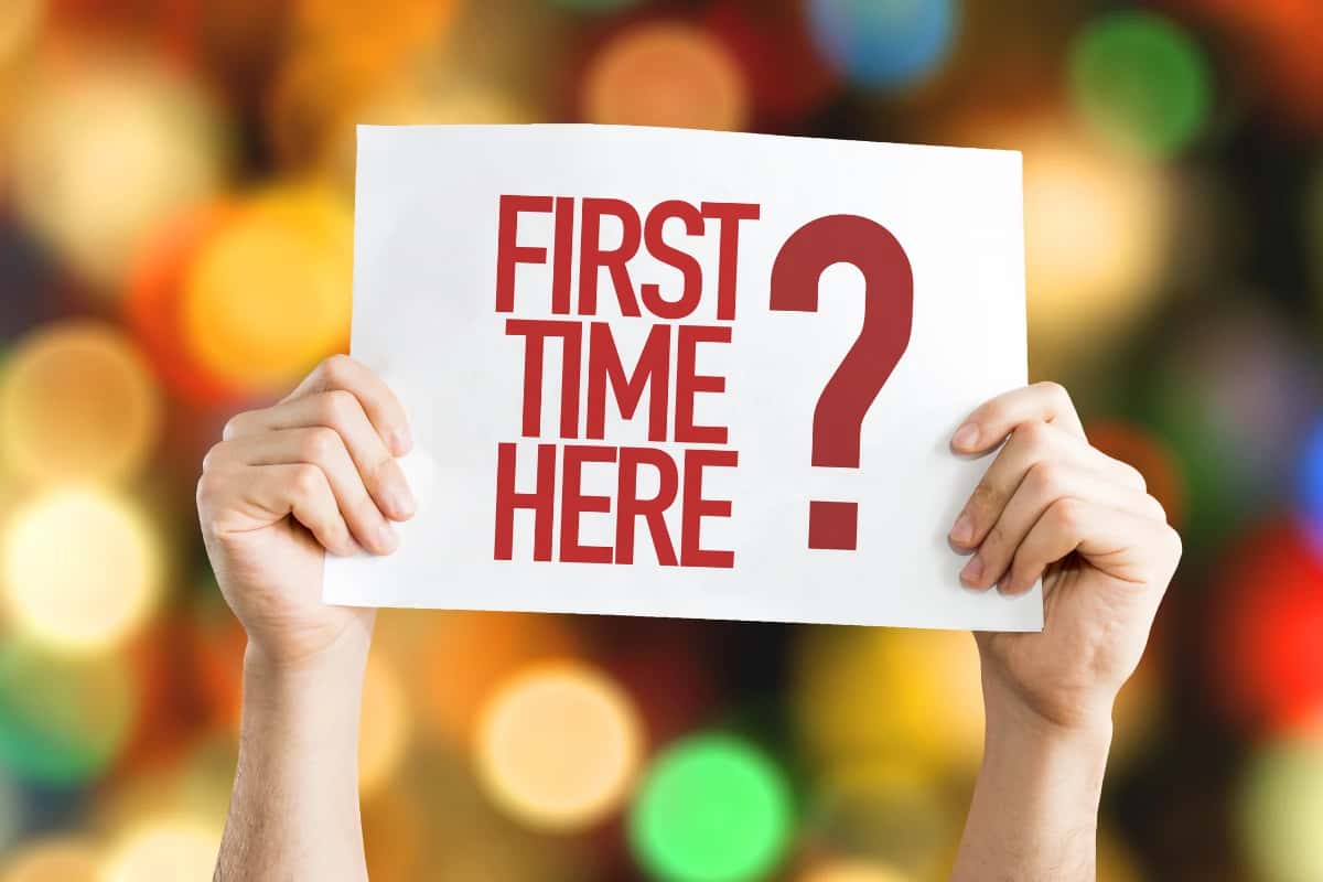 Hands holding a sign reading "FIRST TIME HERE?" against a colorful, blurred background.