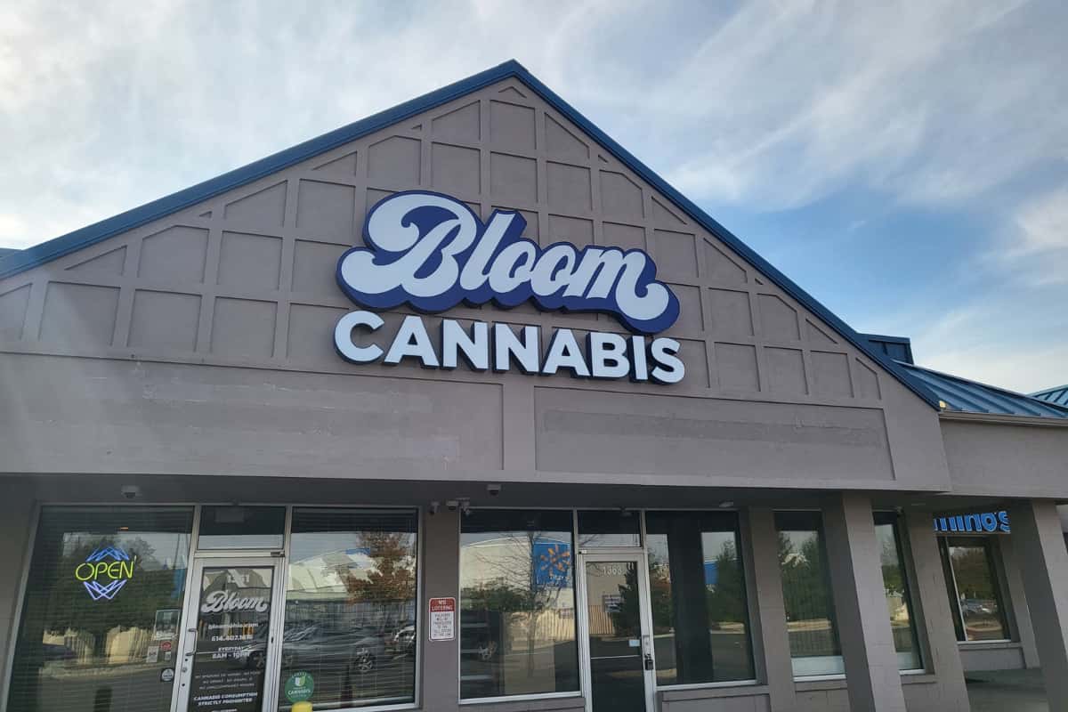 Front of Bloom Cannabis storefront with large blue logo on gray gabled facade and an "Open" neon sign in the window.