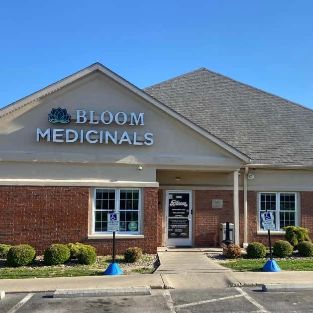 Bloom Medicinals storefront with lotus logo on beige brick building, central glass door, walkway and two handicap parking signs under clear blue sky.