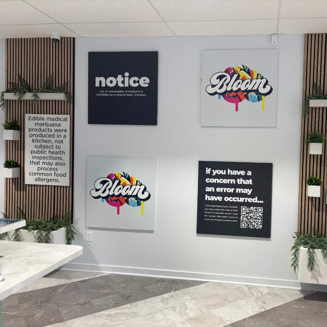 Bloom south columbus interior dispensary wall with four framed signs: two say "Bloom" with colorful designs, one is an error notice with a QR code, and one is about edible marijuana products.