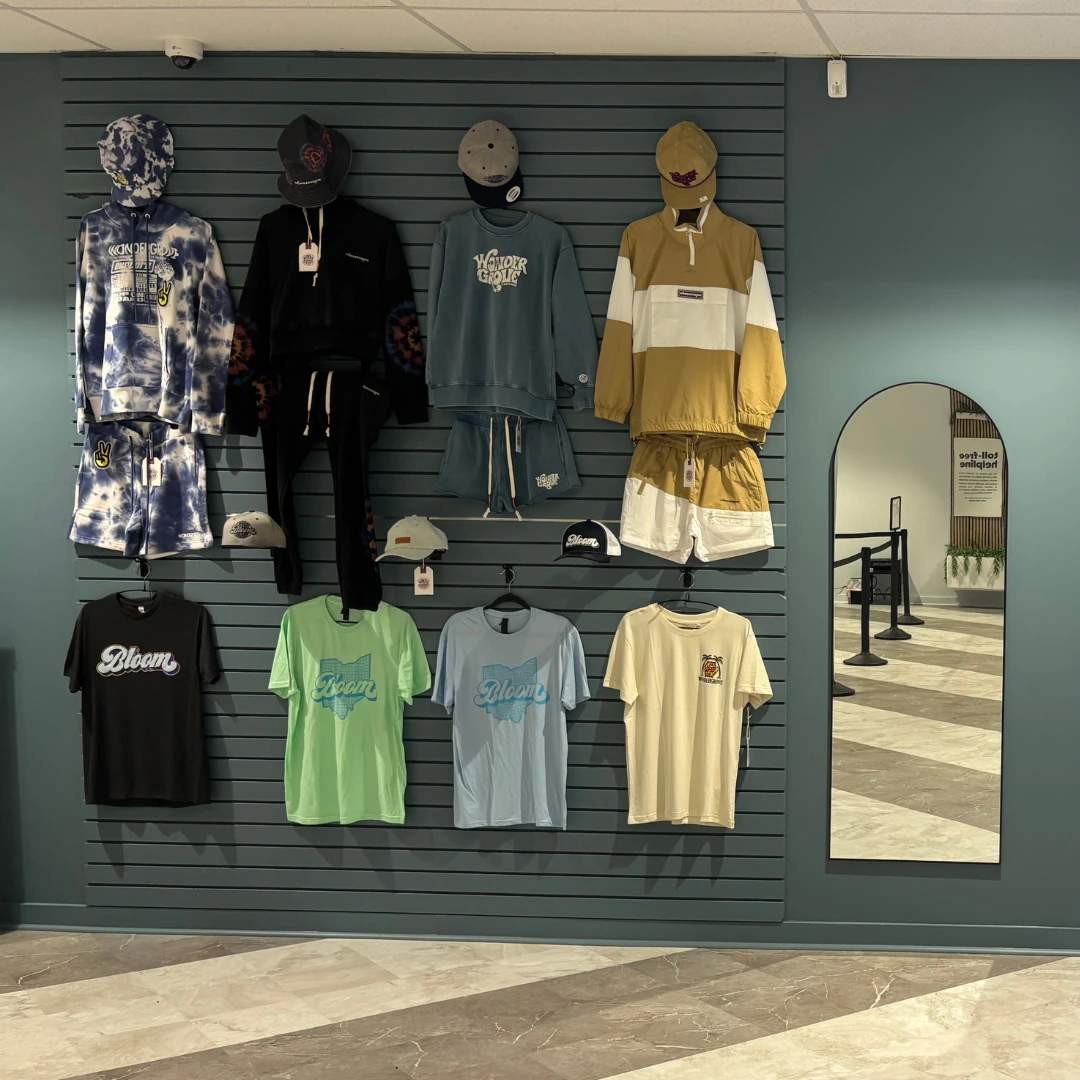 Bloom south columbus dispensary wall with clothing display on a teal wall with various shirts, shorts, and hats. There is a tall mirror on the right and a rope barrier in the background.