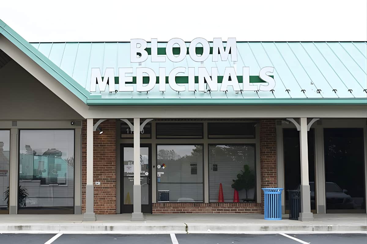 Storefront of Bloom Medicinals with large white sign on teal metal roof, brick facade, empty windows and a blue trash can.