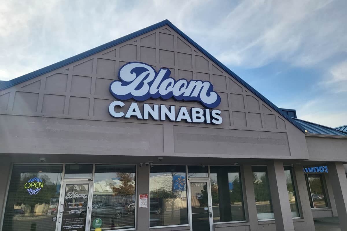 Front of Bloom Cannabis storefront with large blue logo on gray gabled facade and an "Open" neon sign in the window.