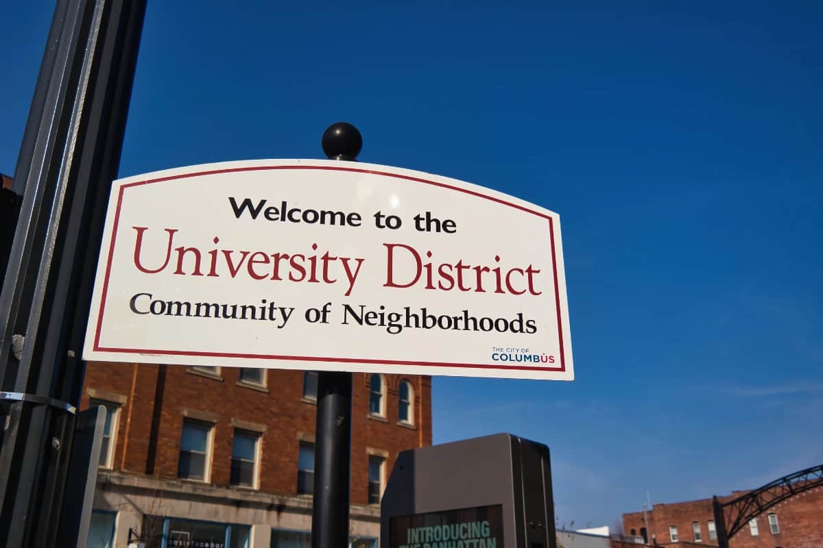 Sign reading “Welcome to the University District, Community of Neighborhoods” in Columbus, Ohio against a clear blue sky and brick buildings.
