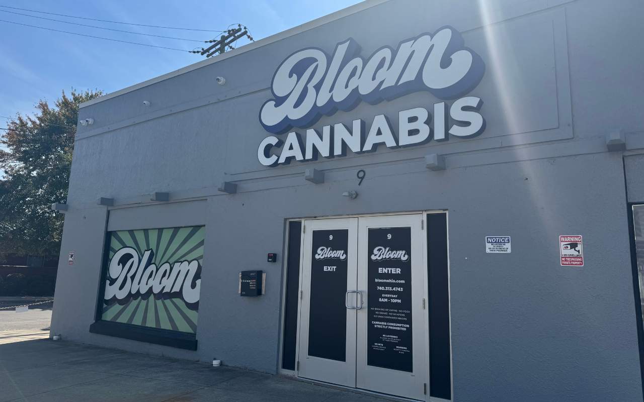 Storefront of Bloom Cannabis with large signage, gray exterior, and entrance doors. Sunlight casts shadows on the building.