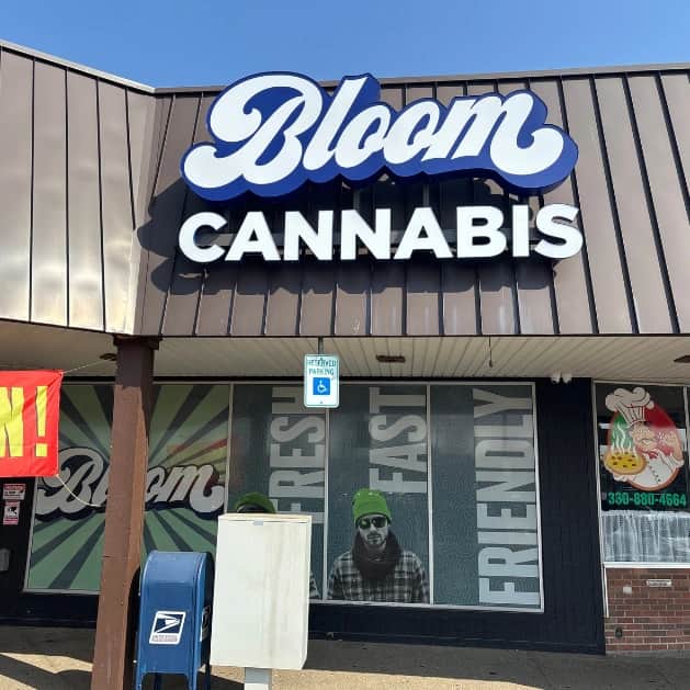 Bloom Cannabis storefront with large blue Bloom sign, window graphic of man in green beanie, handicap parking sign and mailbox