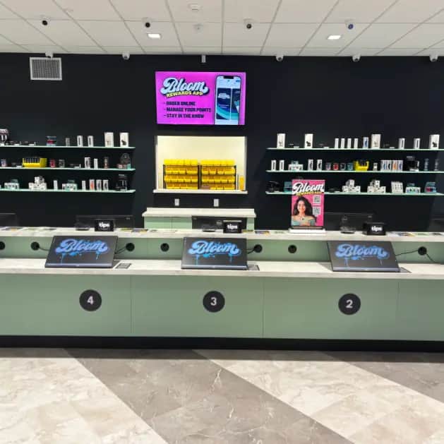 Modern dispensary checkout with numbered counters, Bloom touchscreen displays, product shelves and a pink rewards-app ad on a black wall.