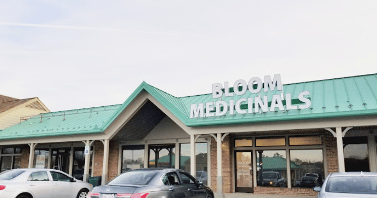 Storefront of Bloom Medicinals with a green roof, brick exterior, and parked cars in front.