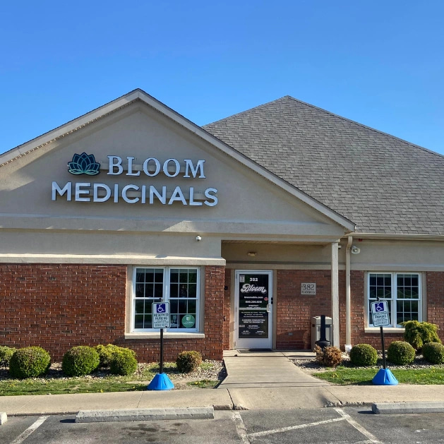 Bloom Medicinals storefront with lotus logo on beige brick building, central glass door, walkway and two handicap parking signs under clear blue sky.